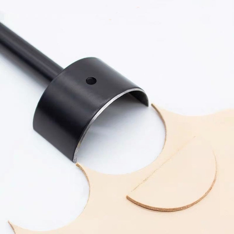 May include: A black leathercraft tool with a cylindrical head and a long handle. The tool is positioned over a piece of light tan leather, with a curved cut visible. The tool is designed for cutting leather.