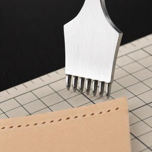 May include: A stainless steel leather stitching chisel tool with multiple prongs is shown next to a piece of tan leather with evenly spaced stitch holes. The tool is used for creating holes for hand stitching leather projects.