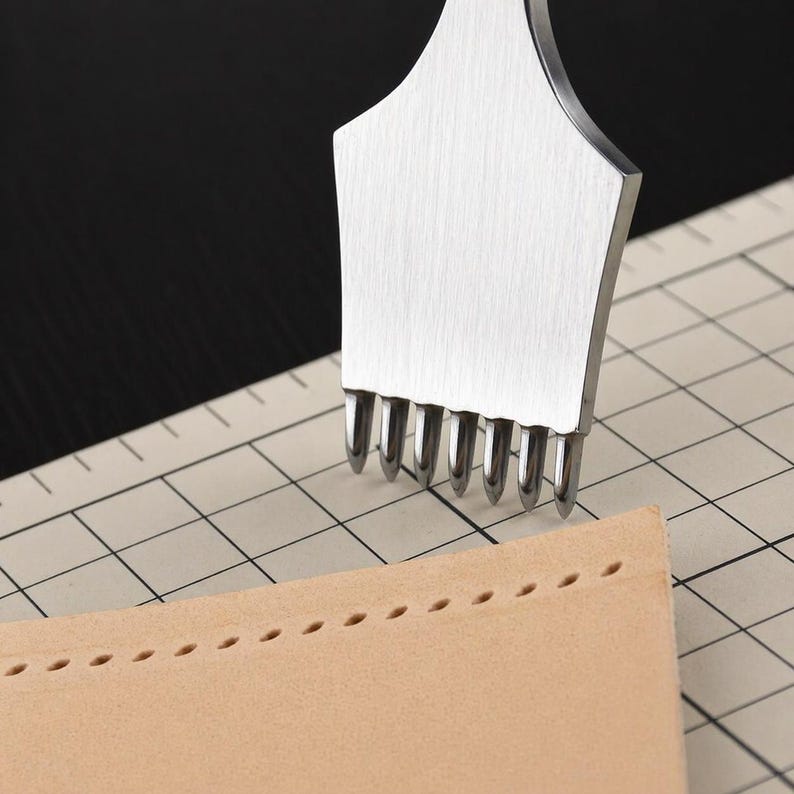 May include: A stainless steel leather stitching chisel tool with multiple prongs is shown next to a piece of tan leather with evenly spaced stitch holes. The tool is used for creating holes for hand stitching leather projects.
