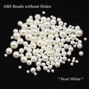 Op de afbeelding: Een verzameling witte, ronde ABS-kralen zonder gaten, in verschillende maten, verspreid op een zwart oppervlak. De tekst "ABS Beads without Holes" en "* Pearl White *" zijn zichtbaar.