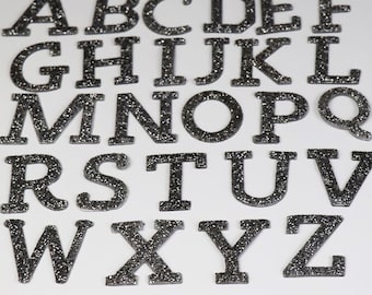 Peut inclure: Un ensemble de 26 lettres en paillettes argentées, de A à Z, dans un style de police en bloc. Les lettres sont disposées en trois rangées sur un fond blanc.