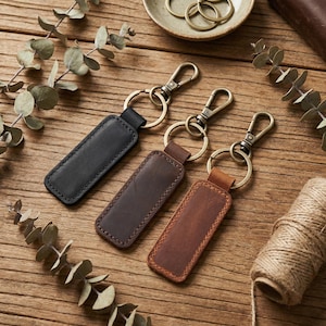May include: Three leather keyrings in black, brown, and tan. Each keyring features a rectangular leather tag with a metal clasp and ring. The keyrings are displayed on a wooden surface with sprigs of greenery.