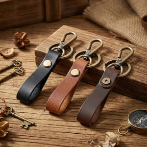May include: Three leather keyrings in black, tan, and dark brown, each with a brass-coloured clasp and a rivet detail. The keyrings are displayed on a wooden surface, with antique keys and a compass in the background.