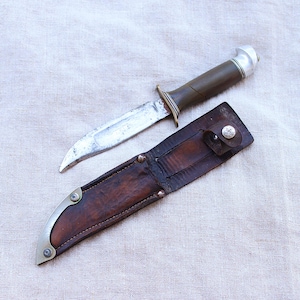 Op de afbeelding: Een vaststaand mes met een zilverkleurig metalen lemmet, een donker olijfgroen handvat en een zilveren pommel. Het mes wordt vergezeld door een donkerbruine lederen schede met een metalen punt, decoratieve klinknagels en een drukknoopsluiting.