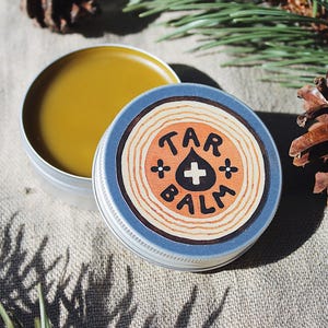 Può includere: Un barattolo aperto color argento con balsamo giallo accanto a un barattolo chiuso con bordo blu e un disegno a venature di legno. Il coperchio del barattolo chiuso presenta le parole "TAR BALM" con un simbolo a goccia e croce.
