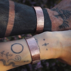 May include: Two copper-colored, textured metal bracelets. The bracelets are worn on wrists with visible tattoos. The bracelets have a series of vertical lines etched into the metal, creating a ribbed effect. The bracelets appear to be handmade.