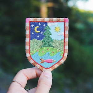 Peut inclure: Écusson brodé avec une scène de nature. Il représente un ciel nocturne avec un croissant de lune et des étoiles, un pin vert, un soleil jaune et un poisson rose dans un lac bleu. Bordure à motif marron et rose.