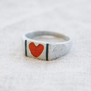 Può includere: Un anello color argento con una faccia rettangolare piatta caratterizzata da un'icona a cuore arancione centrale affiancata da due linee verticali verde scuro. Il lato della fascia dell'anello è inciso con il numero 42 in un font testurizzato.