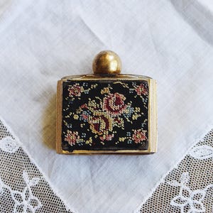 May include: A vintage perfume bottle with a gold-colored cap and trim. The black body is embroidered with a floral design in shades of red, pink, yellow, and blue. The bottle is rectangular and sits on a white surface.
