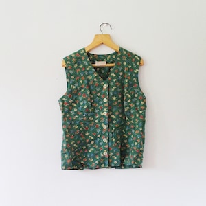 Puede incluir: Un chaleco sin mangas de tela verde oscuro con un estampado floral multicolor de pequeñas flores rojas, amarillas y azules. La prenda tiene un cuello en V, un cierre frontal con cinco botones de color claro y cuelga de una percha de madera.
