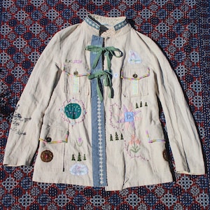 May include: A cream-coloured long-sleeve jacket featuring a mandarin collar, a central blue fabric panel with green ribbon ties, and various embroidered motifs including trees, flowers, a star, and the text E.K. 20217 above a chest pocket.