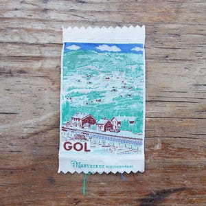 Puede incluir: Un parche rectangular de tela tejida que presenta un paisaje de montañas verdes, un cielo azul con nubes blancas y edificios rojos. El texto GOL está impreso en rojo y el texto AS NARVESENS KIOSKKOMPANI está impreso en azul verdoso abajo.
