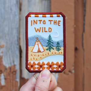 Könnte beinhalten: Ein gestickter Aufnäher mit rotem, orangem und weißem Rand. Der Aufnäher zeigt einen blauen Himmel mit einer Mondsichel, einem kleinen Zelt und zwei Kiefern. Der Text "INTO THE WILD" ist in orangem Garn gestickt.
