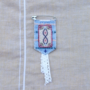 Peut inclure: Une broche textile faite à la main avec un fond bleu clair, présentant un motif brodé avec des accents rouges, blancs et bleus. Une épingle de sûreté argentée fixe le textile, et une gland en dentelle blanche pend du bas. La broche est sur un tissu beige.