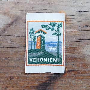 Puede incluir: Un parche de tela tejido rectangular que presenta una escena de paisaje con una torre alta de color naranja y verde, pinos verdes y un cuerpo de agua azul. La palabra VEHONIEMI está bordada en letras mayúsculas blancas en la parte inferior.