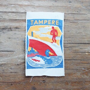 Puede incluir: Un parche bordado rectangular con la palabra TAMPERE en letras amarillas en la parte superior. El diseño incluye una silueta roja de una persona, una gran forma roja curva y un pato azul y blanco nadando en el agua bajo un cielo amarillo.