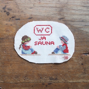 May include: An oval-shaped white cross-stitch fabric piece featuring red text that reads WC JA SAUNA. The design includes two figures sitting in profile, one with a green headband and red shirt, the other with a blue hat and red vest, with -98 in red.
