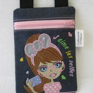 Pode incluir: Bolsa crossbody de ganga com zíper rosa e design de menina de desenho animado. A menina tem cabelo castanho, óculos e está segurando um telefone. O texto diz "J'aime les selfies". Inclui alças pretas e uma etiqueta branca.