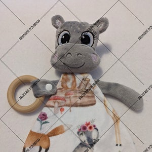 May include: A gray plush hippo-shaped baby comforter with a wooden ring. The comforter features a white fabric with illustrations of a fox, deer, goat, and hedgehog, all adorned with floral crowns. The text "BRODERIE D'ZHIBOUX" is printed on the fabric.