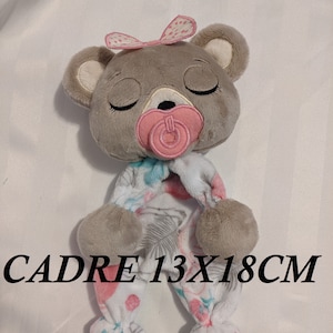 May include: A plush bear doll with closed eyes, a pink pacifier, and a pink bow. The bear is wearing a patterned onesie with blue and pink accents. The text "CADRE 13X18CM" is printed on the image.