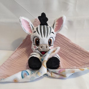 Pode incluir: Um brinquedo de pelúcia de zebra com listras pretas e brancas e orelhas cor-de-rosa está aninhado em um cobertor de musselina para bebê rosa e branco com um padrão de arco-íris.