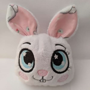 Könnte beinhalten: Ein weißer Plüschhase mit rosa Ohren und gestickten blauen Augen. Die Ohren haben ein weiß-rosa Blumenmuster.