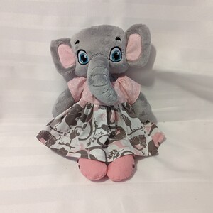 Pode incluir: Um brinquedo de pelúcia de elefante cinza com orelhas, pés rosa e um vestido rosa com bolinhas brancas. O vestido tem um padrão de animais e desenhos florais marrons e rosa. O elefante tem olhos azuis grandes.