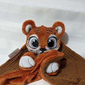 Peut inclure: Une couverture de sécurité en peluche pour bébé en forme de tigre, dans les tons brun et orange. Le tigre a de grands yeux bleus et un museau blanc. La couverture est faite d'un tissu doux et texturé. L'étiquette sur la couverture indique "Félicitations" et "D'zippoux".