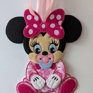 Può includere: Un personaggio di Minnie Mouse in peluche ricamato, con un fiocco rosa e bianco a pois sulla testa, che indossa un body con stampa floreale rosa e bianca e un ciuccio blu. Il personaggio ha orecchie nere e un fiocco rosa. Un nastro rosa pende dalla parte superiore del personaggio.