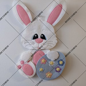 Könnte beinhalten: Ein weißer Filzhase hält ein blaues Filz-Osterei mit bunten Blumen. Der Hase hat rosa Ohren und eine rosa Nase.