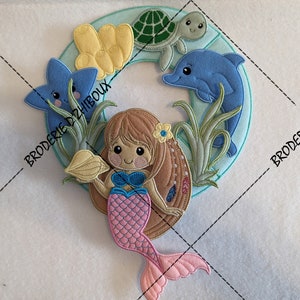 Könnte beinhalten: Ein bunter gestickter Kranz mit einer Meerjungfrau, die eine Muschel hält, einem Delfin, einer Schildkröte und einem Seestern. Der Kranz ist von grünem Seegras und gelber Koralle umgeben.