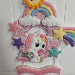 Può includere: Un unicorno di feltro rosa e bianco con un ciuccio in bocca è appeso a un arcobaleno con nuvole e un sole giallo. L'arcobaleno è circondato da stelle rosa e uno stendardo rosa con spazio per il testo.