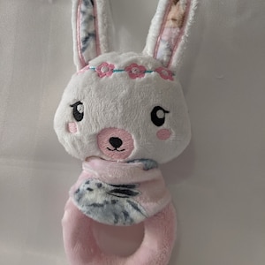 May include: A plush white bunny rattle with pink accents. The bunny has embroidered facial features, a pink flower crown, and patterned ears. The rattle features a pink ring and a patterned neck wrap.