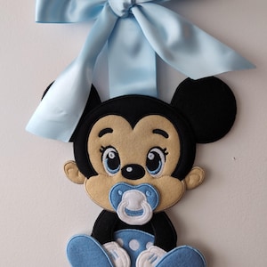 Può includere: Decorazione in feltro di Baby Mickey Mouse con ciuccio, fiocco e dettagli azzurri. Il design nero, beige e blu presenta il testo "Broderie D'Zhiboux" in basso. Progettato per essere appeso.