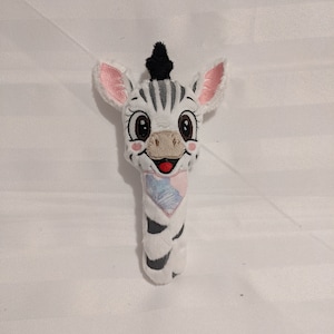 以下が含まれることがあります： 黒と白のシマウマのぬいぐるみで、ピンクの耳と青とピンクの模様のスカーフをしています。おもちゃは、黒い目と赤い口で笑顔をしています。