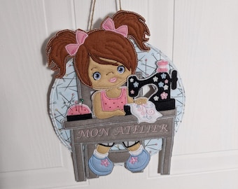 May include: A felt wall hanging featuring a cartoon girl with brown hair and pink bows, seated at a sewing table. The table has the text "MON ATELIER". A sewing machine and pincushion are also depicted. The background is light blue with sewing-themed patterns.