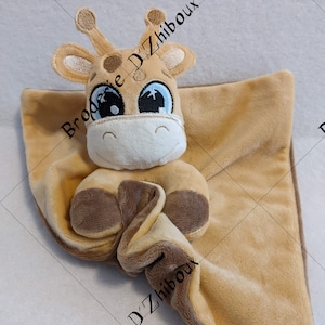 May include: A tan plush giraffe security blanket with embroidered brown spots and large blue eyes. The giraffe is holding the blanket in its paws. The blanket has the text "Broderie D'Zhiboux" embroidered on it.