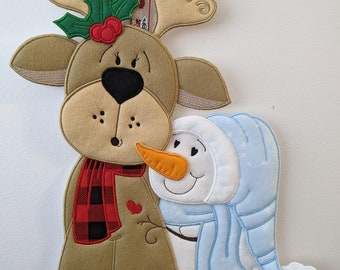 May include: A brown reindeer with red and black plaid scarf and a green holly sprig on its antlers is hugging a white snowman with a blue scarf and hat. The snowman has a carrot nose and black eyes.