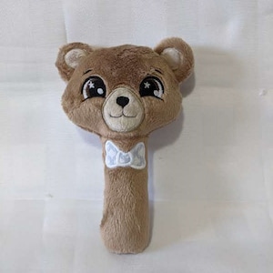 May include: A brown plush bear rattle with a white bow tie. The bear has large, black eyes with white stars inside. The rattle is designed for babies and toddlers.