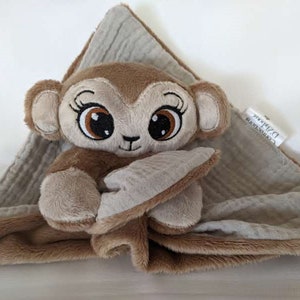 Op de afbeelding: Een bruine pluche aap met grote ogen en een zacht, grijs en wit muslin deken. Het deken heeft een label met de tekst "Comfy Cozy D. Zahara".