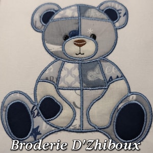 May include: Embroidered teddy bear in shades of blue and grey, with a patchwork design. The bear has a dark brown nose and black eyes, and is sitting with its legs outstretched. The text "Broderie D'Zhiboux" is at the bottom.