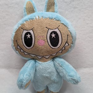 May include: A light blue bunny plush toy with a tan face and a stitched mouth. The bunny has large black eyes with white star accents, a pink nose, and is wearing a hood with bunny ears. The plush toy is made of soft material.