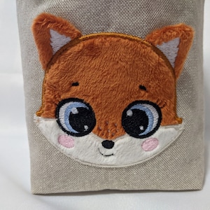 Könnte beinhalten: Ein beigefarbener Stoffbeutel mit einem gestickten Fuchgesicht. Der Fuchs hat orangefarbenes Fell, große blaue Augen und rosa Wangen. Der Beutel ist rechteckig und scheint aus einem strukturierten Stoff zu bestehen.
