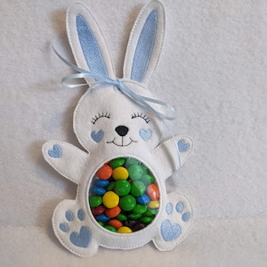 Puede incluir: Un conejito de fieltro blanco con orejas azules y una ventana de plástico transparente en el vientre. La ventana está llena de caramelos de colores. El conejito tiene una cinta azul atada alrededor del cuello.