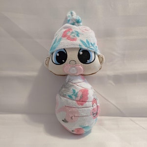 May include: A plush baby doll toy wrapped in a pink and white blanket with a floral pattern. The doll is wearing a blue and white hat with a floral pattern. The doll has large blue eyes and a pink pacifier.