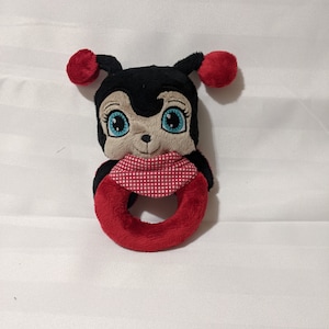 Puede incluir: Un juguete de sonajero de peluche en forma de mariquita con una bufanda a cuadros rojos y blancos, un cuerpo negro y grandes ojos azules.