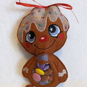 May include: A brown felt gingerbread man-shaped pouch with embroidered details. The pouch has a clear window filled with colorful candies. It features a red bow and is decorated with white frosting and sprinkles.