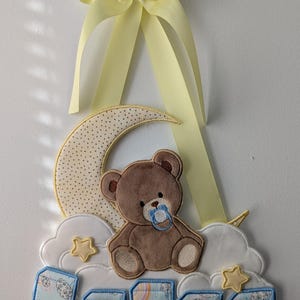 May include: A baby nursery decoration featuring a brown teddy bear sitting on a yellow crescent moon with white clouds and stars. The moon is decorated with yellow polka dots. The decoration is hung with a yellow ribbon bow.