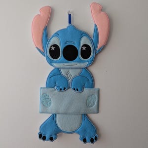 May include: Blue and pink felt Stitch character wall hanging with a blank white felt sign. The character has large blue eyes with white stars and a blue mouth.