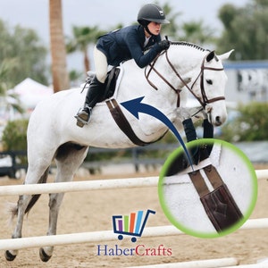 May include: A white horse jumping over a white fence with a rider wearing a black jacket and a helmet. The horse is wearing a brown leather saddle and a brown leather girth with a white stripe.